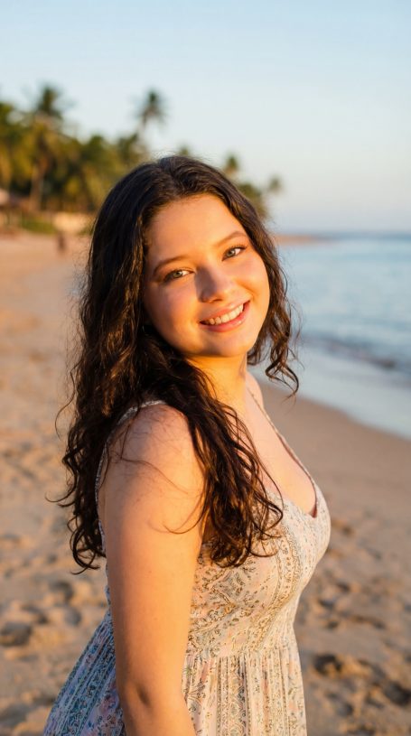 Ellie Fontaine Junge Frau mit langen, lockigen Haaren lächelt am Strand bei Sonnenuntergang.