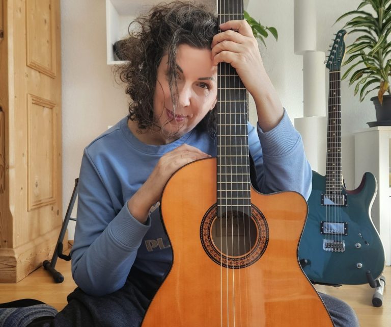 © Christien Marie Wach Christien sitzt mit akustischer Gitarre, Gesicht leicht zugewandt und fröhlich.